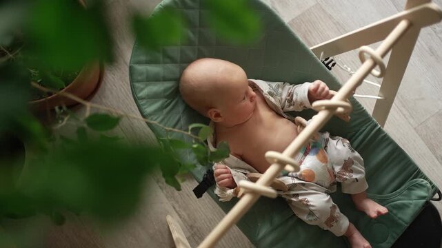 Curious newborn baby resting in a soft, green bouncer seat with wooden toys, looking around the room from a top down perspective while enjoying a moment of quiet wakefulness at home