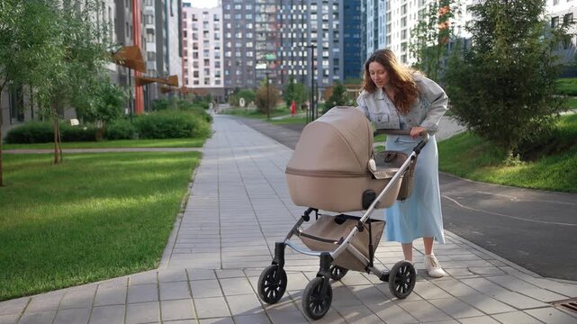 Caring mother pushes her baby's stroller along a sidewalk in a contemporary urban neighborhood, stopping to affectionately check on her child while on a pleasant walk together