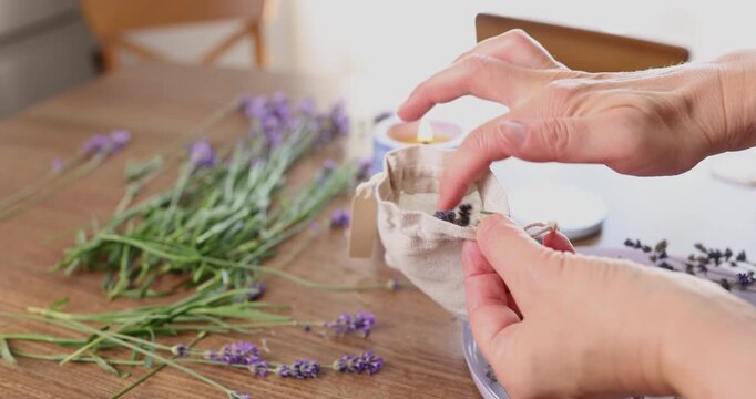 Woman hands putting dried lavender flowers inside small cotton sachet. Creative process making handmade scented pouch on wooden table workspace. Aromatherapy craft hobby diy home decor concept video.