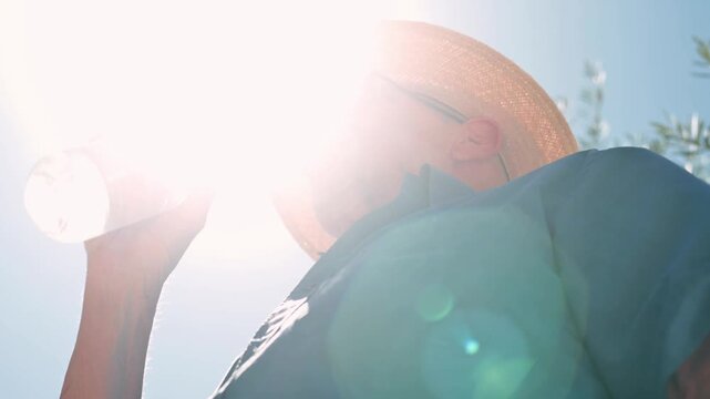 A man in a straw hat drinks from a water bottle under intense sunlight, with pronounced lens flare and blue sky suggesting summer heat, refreshment, outdoor leisure and cooling relief.
