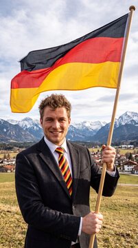 A smiling man in a dark suit and a german flag tie holds a large german flag on a wooden pole, standing in a grassy field with snow-capped mountains and a village in the background