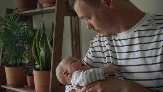 Young caucasian father gently holding his infant son in his arms, rocking and comforting the baby in a cozy room with houseplants, embodying a warm moment of paternal love and family bonding