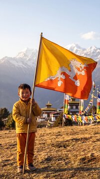 A young bhutanese boy wearing a yellow jacket stands in a grassy field, holding a large bhutan flag with a white dragon, against a backdrop, of snow-capped himalayan mountains
