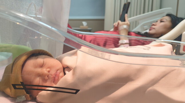 Newborn Sleeping in Hospital Bassinet with Mother Resting in Background