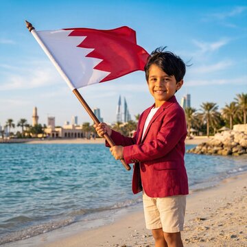 A young boy wearing a maroon blazer and beige shorts stands on a sandy beach, holding a waving flag of bahrain against a backdrop, of the manama, city skyline and, blue ocean water