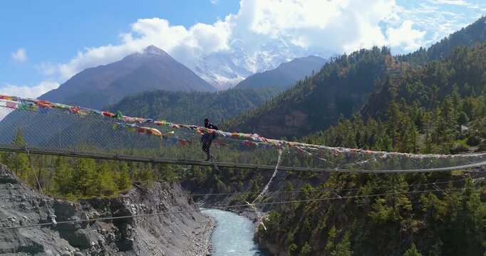 Annapurna Circuit, Manang Nepal, aerial gliding shot of a trekker crossing a long suspension bridge adorned with prayer flags, suspended high above the Marsyangdi River, mountains in the background