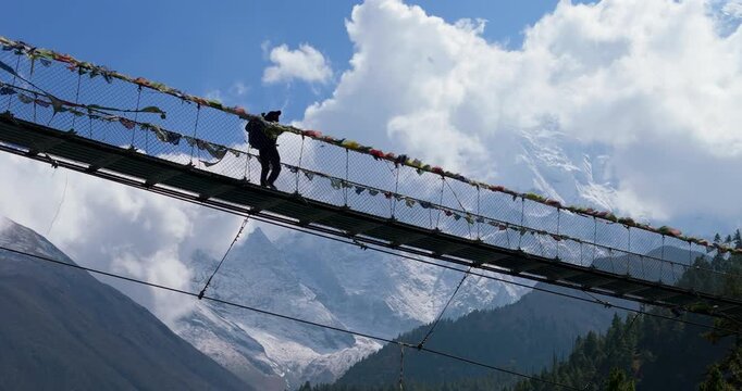 Annapurna Circuit, Manang, Nepal, low-angle tracking shot of a male trekker walking across a long suspension bridge lined with prayer flags above a winding river valley, surrounded by mountains