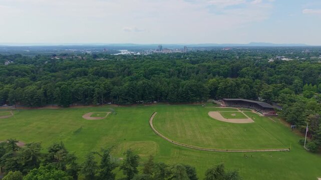 Forward drone fly over expansive green recreation baseball fields bordered by dense woodland with distant Springfield town, Massachusetts, US