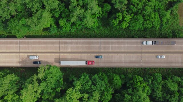 Top-down aerial of a flatbed semi hauling a tarped load across an elevated interstate bridge through dense green forest, with dry van and passenger traffic flowing in both directions.