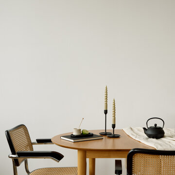 Minimalist dining table with yellow flowers in glass vase, candles, black teapot, and limes, set against a white wall. Copy space.