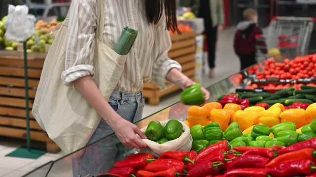 Woman picking fresh green bell peppers from supermarket shelf using reusable ecobag zero waste concept.