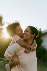 Fototapeta premium Laughing woman giving a piggyback ride to a small child in a sunlit backyard, featuring hazy summer light and soft ribbed textures for family bond and parenting lifestyle content