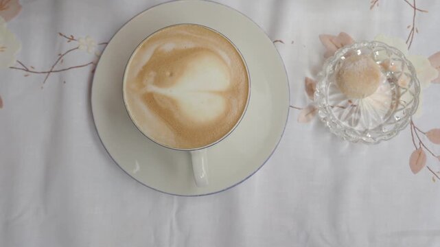 Top-down view of a steaming cup of cappuccino adorned with heart-shaped latte art and a sweet biscuit in a glass bowl on a white floral tablecloth
