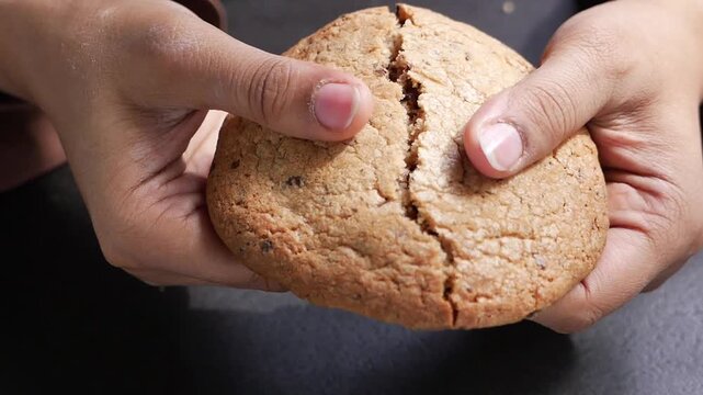 Close up slow motion video of hands breaking fresh homemade chocolate chip cookie in half, revealing gooey inside, delicious dessert snack