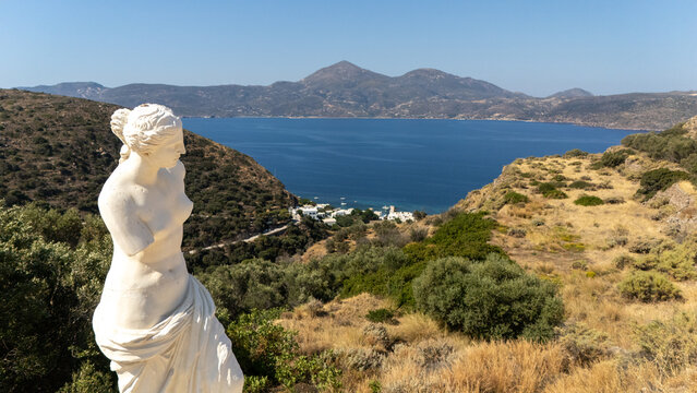 Marble statue of Venus de Milo facing the scenic bay and mountains of Milos, Greece, under clear summer skies.