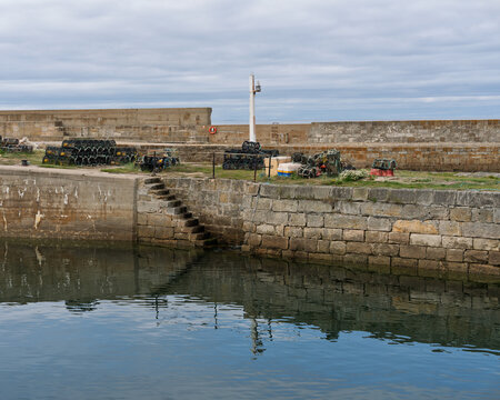 Stone stairs leading into the calm waters of Lossiemouth Harbour in Moray during a cloudy afternoon with fishing equipment stacked on the pier