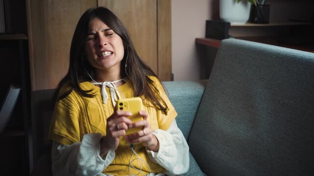 Young Hispanic woman sitting on her sofa at home, wearing headphones, happily singing along to her favorite music while enjoying a joyful and relaxing moment in a cozy home environment.
