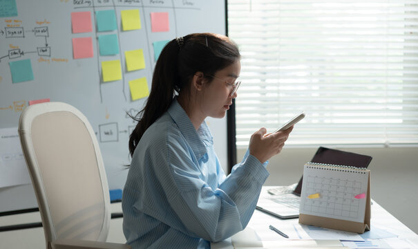 Businesswoman using smartphone while reviewing project calendar and workflow board in a modern office, managing business planning, scheduling, communication and time management.