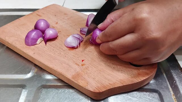 Close-up of a person's hand slicing fresh shallots with a kitchen knife on a wooden cutting board. Food preparation, cooking ingredients, home kitchen, healthy meal concept.