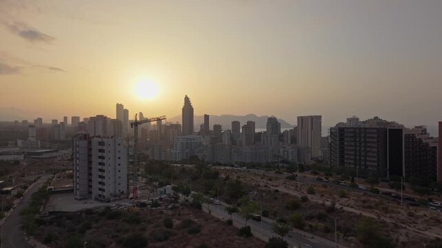 The sun sets over the city skyline as buildings stand tall. The sky shows orange and yellow colors, marking the end of the day during late afternoon hours.
