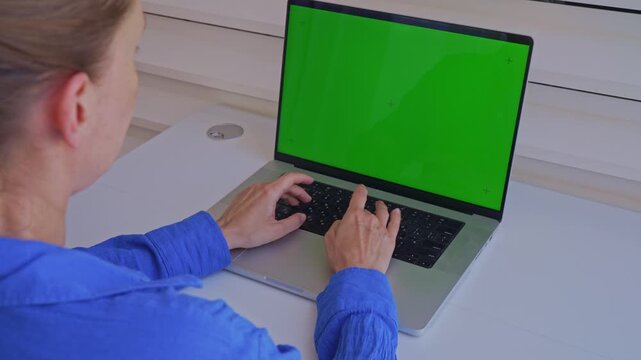 A person types on a laptop with a green screen. The desk is simple and bright. We see the person's hands and the screen from different angles. This shows typing activity.