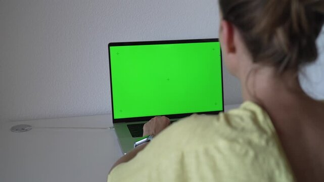 A person sits at a desk and types on a laptop with a green screen. The setting looks like a home office. It is daytime and the person focuses on their work.