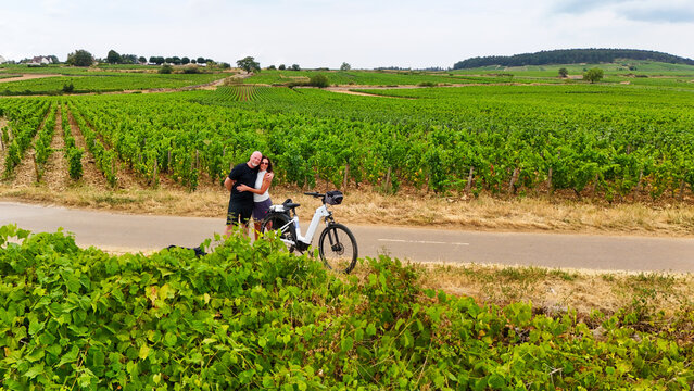 A couple stands beside an electric bicycle on a country road surrounded by lush chardonnay vineyards in Burgundy, France, capturing romance and rural wine‑country charm.