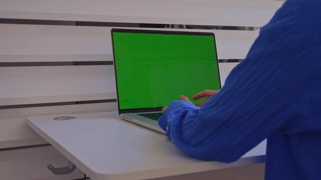 A person sits at a desk and types on a laptop. The laptop screen shows a green background. The setting is indoors with plain walls. The activity focuses on typing.