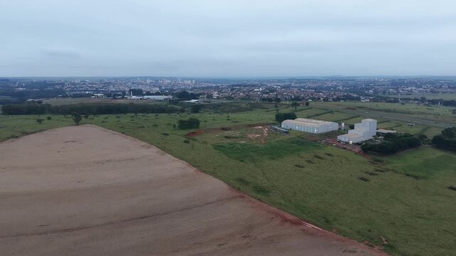 Aerial view of agricultural land and industrial buildings near a city skyline under a cloudy sky