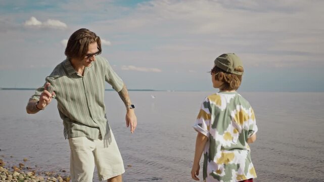 Caucasian father and young son standing on pebble beach, throwing skipping stones into calm sea. Clip is perfect for family travel and outdoor lifestyle commercials