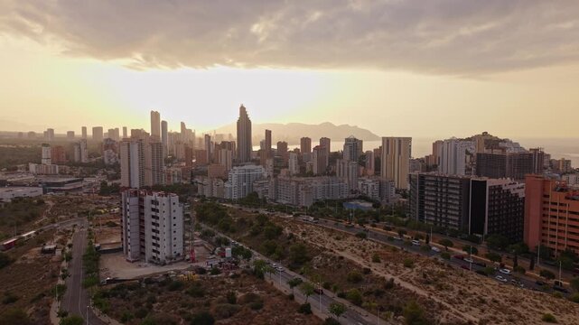 The view shows a city skyline at sunset. Buildings fill the view while the sky changes colors. Streets appear busy with some activity. The scene captures the end of the day.