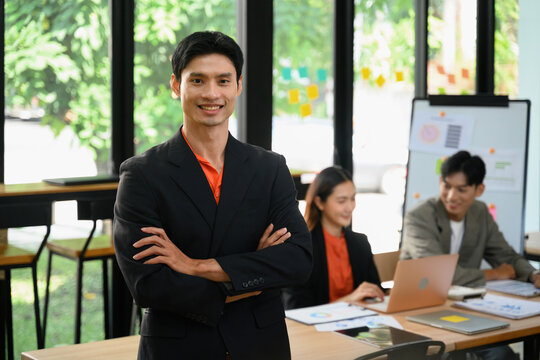 Confident Asian Business Leader Standing Arms Crossed in Modern Office with Team Working in Background