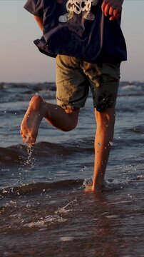 close up of boy running barefoot on beach coast at sunset, sea water is splashing. Carefree childhood. slow motion