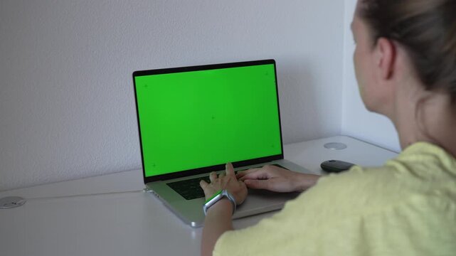 A person sits at a table and works on a laptop with a green screen. The person types and interacts with the device in a bright and simple room.