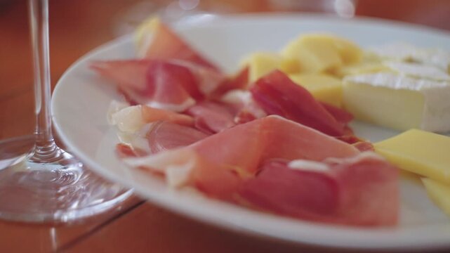 Cheese, meat ham in plate and glass of wine on wooden table in cafe, close-up.