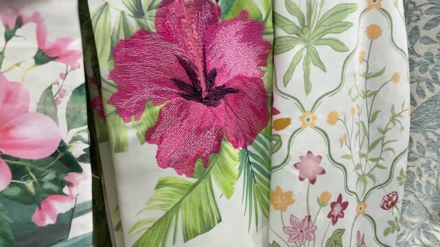 Fabrics closeup. Bright floral fabric display with tropical pink hibiscus prints and embroidered foliage draped on retail shelf. Textile close up. Video taken on mobile device.