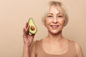 Smiling mature woman holds half avocado near face on beige background. Concept of healthy...