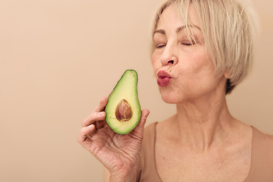Mature woman with short blonde hair holds avocado half close to face, eyes closed, lips puckered as if to kiss fruit. Concept of healthy lifestyle, natural food, wellness, and self-care.