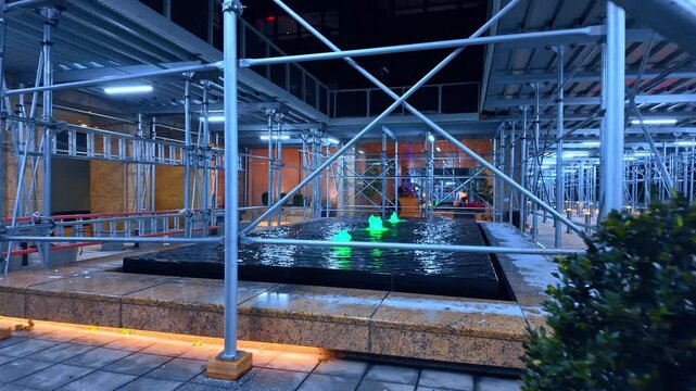 Green fountain under sidewalk scaffolding at Midtown office plaza. Night view shows metal supports surrounding a rectangular water feature with green lights and patches of snow.
