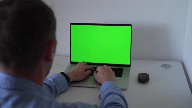 A person sits at a desk typing on a laptop with a green screen display. The setting is a simple home office. The time is afternoon.
