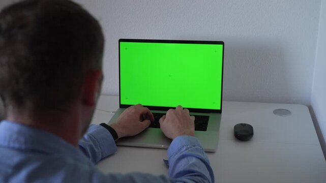 A person types on a laptop that has a green screen displayed. The setup takes place in a home office space. The user appears focused on the work being done.