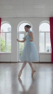 Vertical video. Young elegant ballerina in a blue tutu gracefully performing a classical dance routine on pointe shoes in a spacious, light-filled studio with large windows and a mirror wall