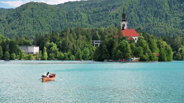 Rowing boat with two unrecognizable people crossing Lake Bled toward Bled Island and the iconic Church of the Assumption in Slovenia on a peaceful summer day.