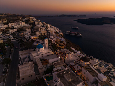 Evening twilight drone aerial landscape of Fira, Santorini, Greece showing traditional white architecture, and cruise ships moored in the Aegean Sea.
