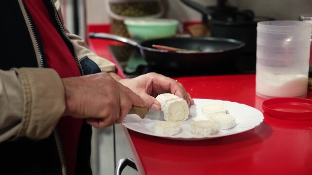Senior male hands slicing a fresh log of goat cheese on a white plate in a modern kitchen