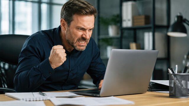 Angry businessman at desk shouting at laptop, clenched fist, papers and office shelves around him