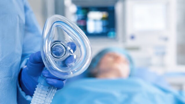 Anesthesia mask held by a gloved hand near a patient in an operating room with monitoring equipment