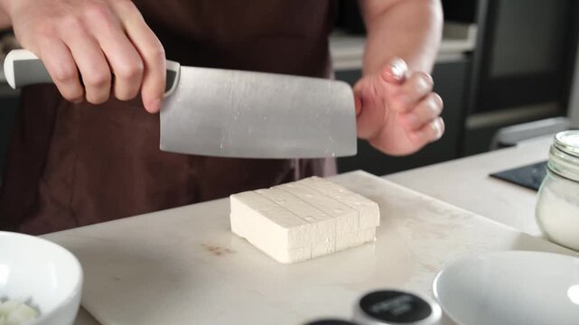 Male cook slicing a block of tofu with a knife on a cutting board in the kitchen