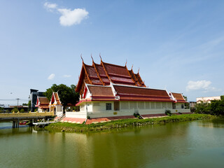 Fototapeta premium Traditional Thai temple building with red roof situated beside a peaceful pond under clear blue sky.
