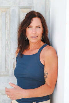 Natural light portrait of a woman with long brown hair wearing a blue tank top, standing against a textured wall with a calm, confident expression.